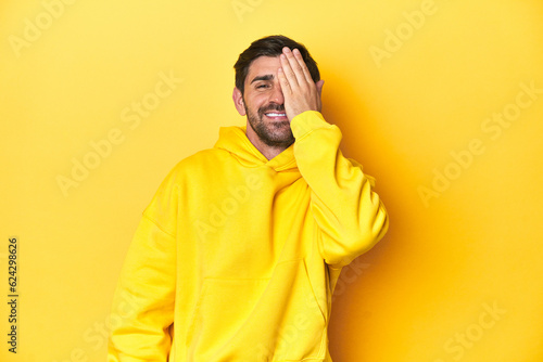 Man in yellow hoodie, monochrome studio backdrop having fun covering half of face with palm.