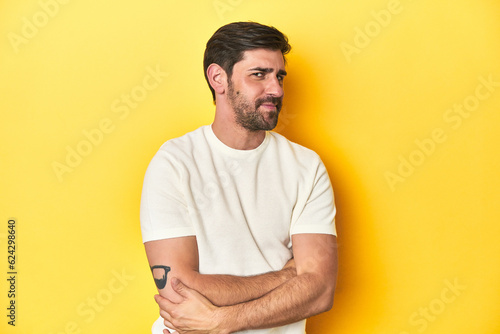 Caucasian man in white t-shirt on yellow studio background suspicious, uncertain, examining you.