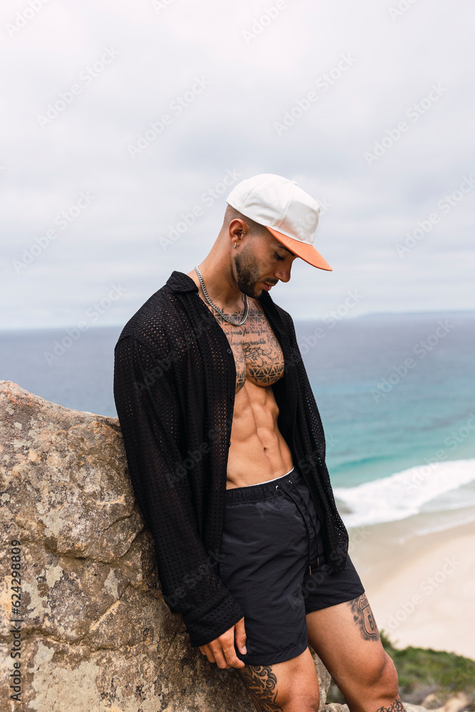 Chico joven tatuado y musculoso posando en acantilado frente a la playa en un día soleado de verano