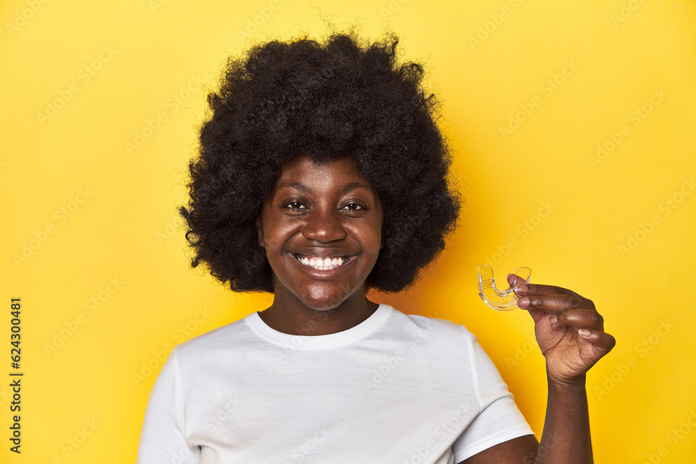 African-American woman holding invisible teeth corrector on a yellow background. African-American woman holding invisible teeth corrector on a yellow background.