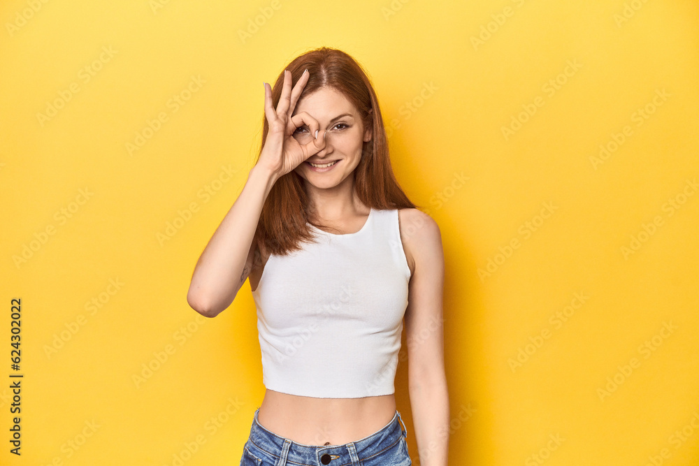 Redhead in white tank top, relaxed studio pose excited keeping ok gesture on eye. Redhead in white tank top, relaxed studio pose excited keeping ok gesture on eye.
