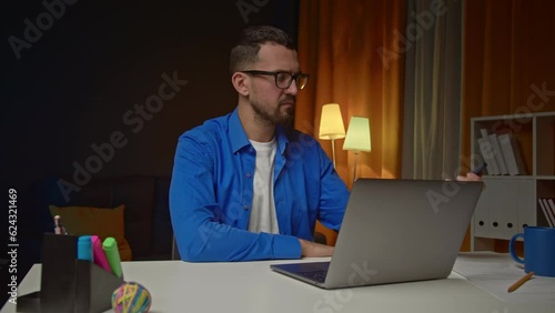 Man using laptop and smartphone and sitting at table in home office. Young freelancer looks at computer screen then takes phone in hands and types, sits at desk in the room.