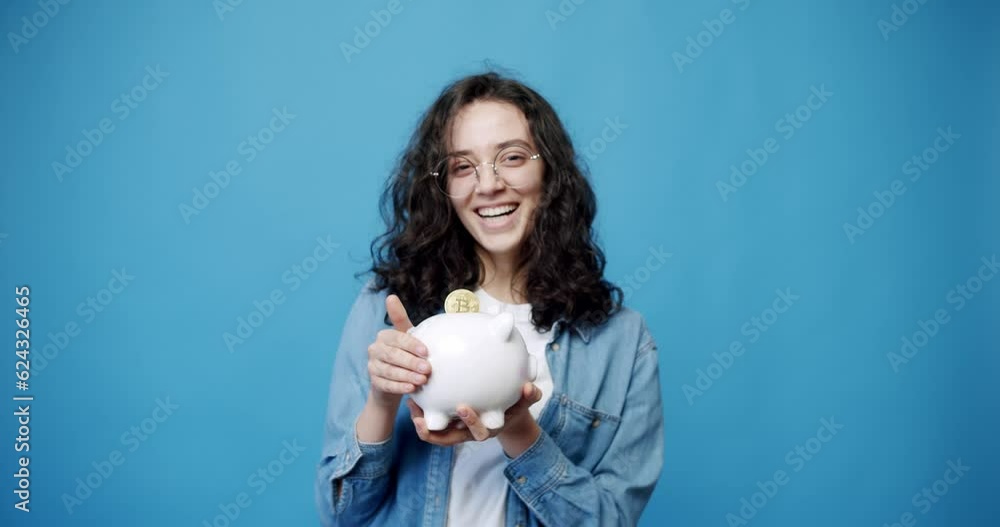 A young woman holds a saving coins box and save money in her hand on isolated blue background. Cute Money Box. Child's Coin Bank. Cute Piggy Bank Money Box. A young woman holds a saving coins box and save money in her hand on isolated blue background. Cute Money Box. Child's Coin Bank. Cute Piggy Bank Money Box.