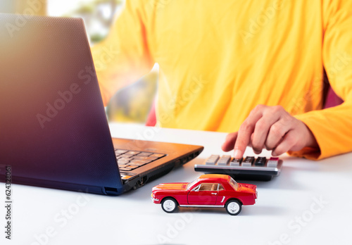 Man using calculator and laptop to find out car loan or tax. Buying new car or insurance concept. Hand pressing calculator with red car in front. 