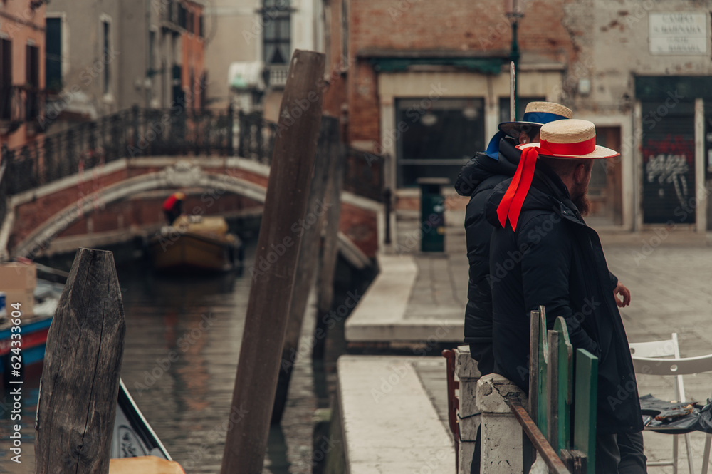 Two gondoliers, dressed in traditional attire, wait by the sidewalk of a Venice canal as they prepare to ferry passengers, with a bridge in the background.