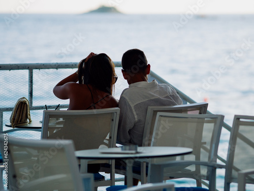 Couple of lovers sits embracing in restaurant against backdrop of sea. Unrecognizable silhouettes dating. Sunset on Adriatic. Cozy resort town in summer. Rovinj, Croatia - July 7, 2023 © klevo