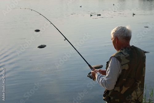 Old man fishing. Senior gray haired fisherman throws a spinning from shoreside at sunset, twisting a coil. Positive elderly male angling at lake, rotating reel rod. Retired fisher enjoying his weekend