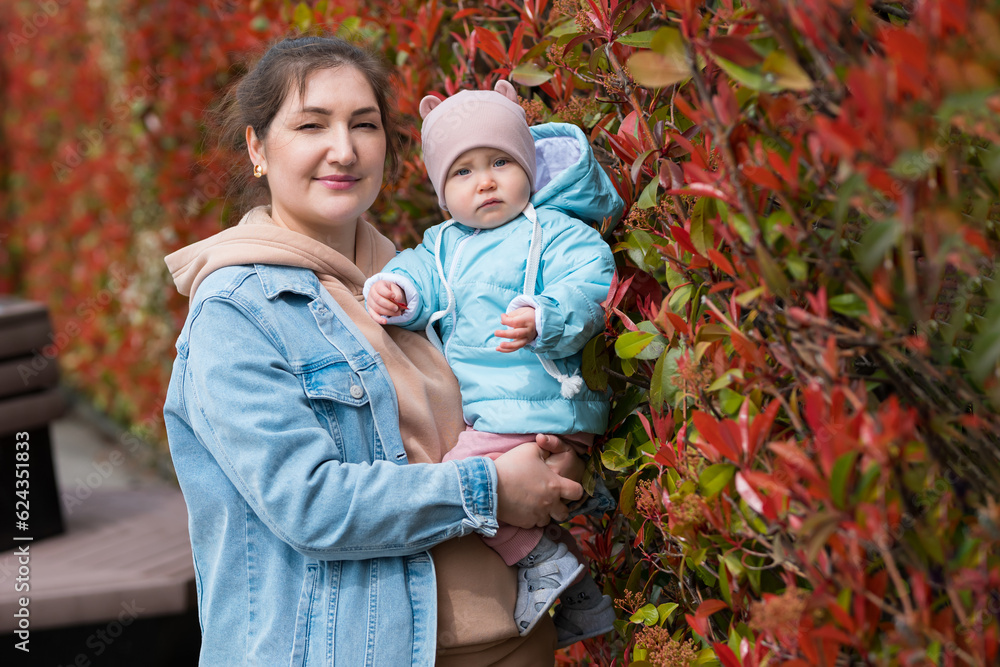 Mother holding in arms toddler daughter in warm clothes posing against bushes with red leaves. Parent and child in similar palette clothes in park Mother holding in arms toddler daughter in warm clothes posing against bushes with red leaves. Parent and child in similar palette clothes in park