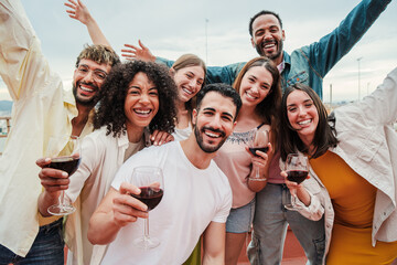 Group of young adult best friends having fun toasting a red wine glasses at rooftop reunion or birthday party, drinking alcohol. Happy people enjoying on a social gathering celebrating together. High
