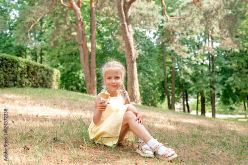 Blonde girl eats ice cream in the park sitting on the grass.