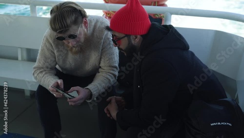 two friends are looking at phone and discussing while they are on the ferry. tourists, bearded men. travel by water