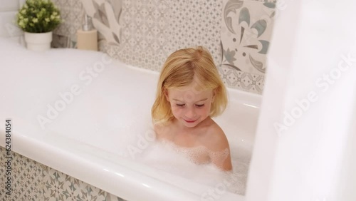 Adorable caucasian three years old gir with curly hair taking bath with white foam bubbles, playing,laughing,having fun.Carefree childhood, kid body care and hygiene concept. photo