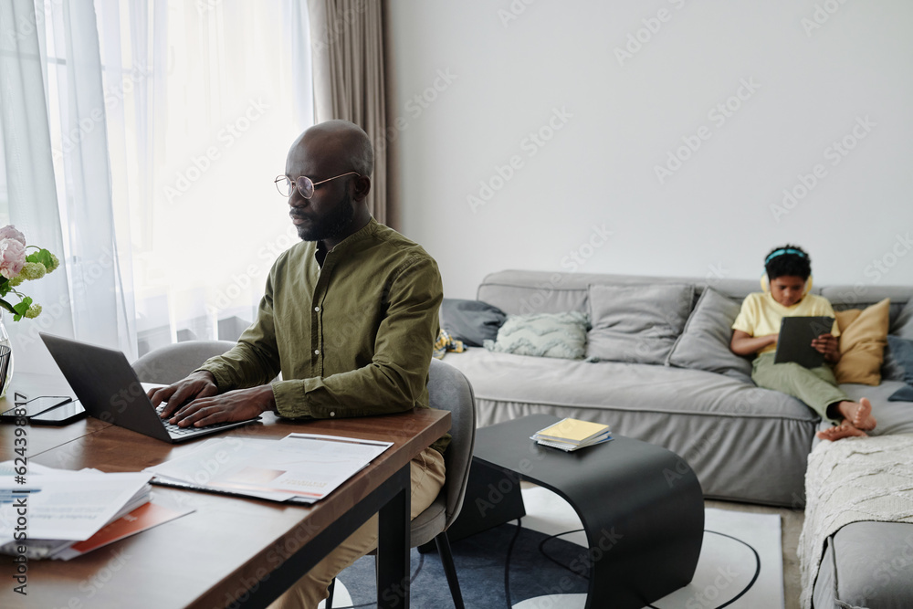 African American dad sitting at table and working on laptop at home while his son playing on tablet pc in background African American dad sitting at table and working on laptop at home while his son playing on tablet pc in background