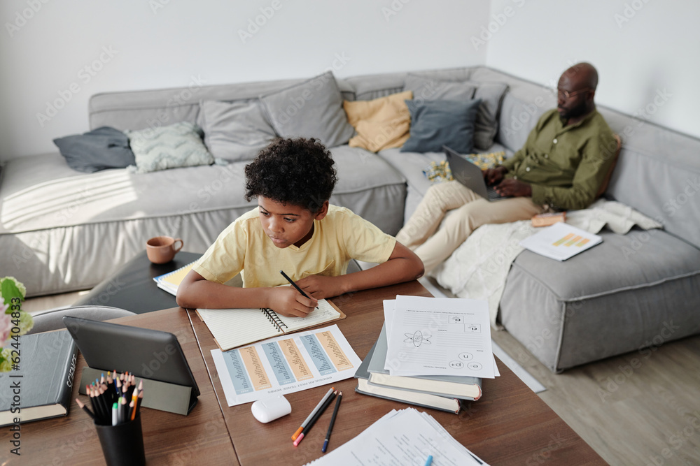 High angle view of African American boy doing homework at table with his dad working on laptop in background High angle view of African American boy doing homework at table with his dad working on laptop in background