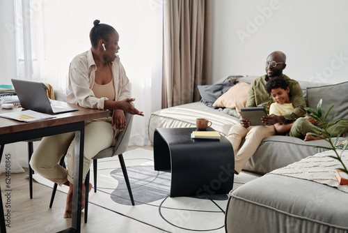 African American mother working online on laptop at home while her family having fun with tablet pc on sofa in background