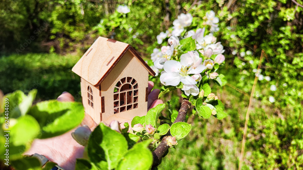 Small wooden toy house on palm of woman hand on natural background with apple flowers. symbol and concept of care, buying, selling, donating of eco friendly home. close-up, soft selective focus