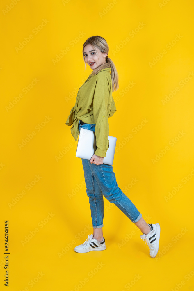 Holding closed laptop, full body side view young blonde woman holding closed laptop. Wearing jeans and green shirt, isolated yellow studio background. Walking, going, IT professional lifestyle concept Holding closed laptop, full body side view young blonde woman holding closed laptop. Wearing jeans and green shirt, isolated yellow studio background. Walking, going, IT professional lifestyle concept