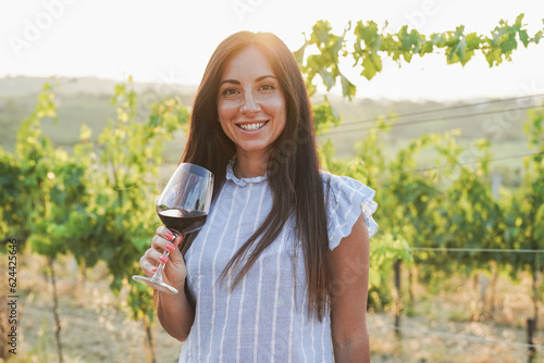 Young woman inside wineyard holding glass of red vine during sunset time and smiling on camera