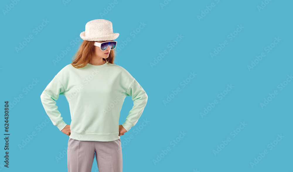 Funny young woman in pastel mint sweat shirt, white faux fur hat and sunglasses looking at copy space side while standing with hands on hips isolated on blue background. Fashion concept Funny young woman in pastel mint sweat shirt, white faux fur hat and sunglasses looking at copy space side while standing with hands on hips isolated on blue background. Fashion concept