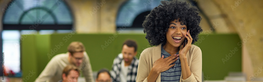 Fototapeta premium Exciting news. Young overjoyed mixed race woman talking on smartphone and smiling while standing in the coworking space with colleagues working on the background