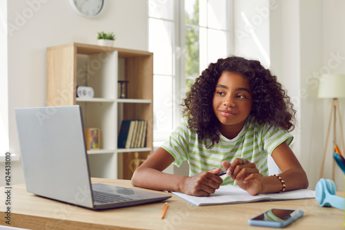 Happy school child having online class. Beautiful African American girl sitting at desk at home, looking at laptop computer, holding pen and smiling. Education, technology, distance learning concept