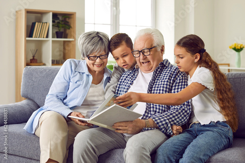 Portrait of a happy grandparents reading a book to their grandchildren or looking through a family photo album sitting on sofa at home. Kids enjoying leisure time with grandma and grandpa on weekend