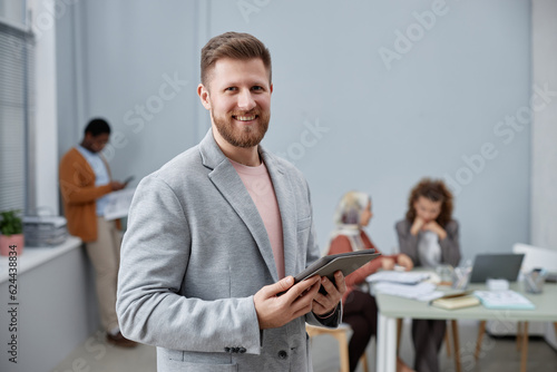 Happy young chief executive officer in elegant grey suit holding tablet while standing in front of camera against coworkers and networking