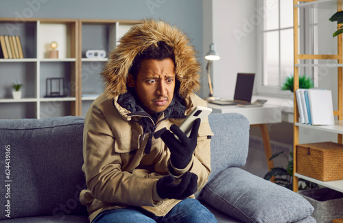 Disappointed young man in warm jacket freezing at home. Unhappy stressed man wearing warm winter clothing feeling cold inside house at cold season. Heating problems, power crisis concept