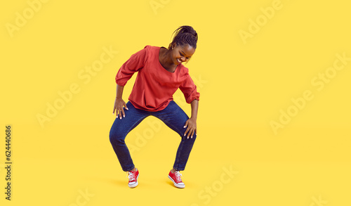 Happy young woman dancing against a vivid vibrant yellow colour studio background. Cheerful beautiful plus size African American dancer girl in comfortable jeans doing lively knee moves to music