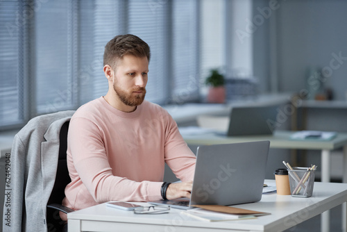 Young serious businessman in casualwear looking through online data on laptop screen and typing while networking or preparing report