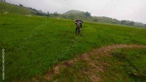 Enduro motorbike biker speed riding extreme stunt tricks wheely on green valley field aerial view. FPV sport drone close shot male racer standing on back wheel motorcycle adrenalin travel adventure 4k photo