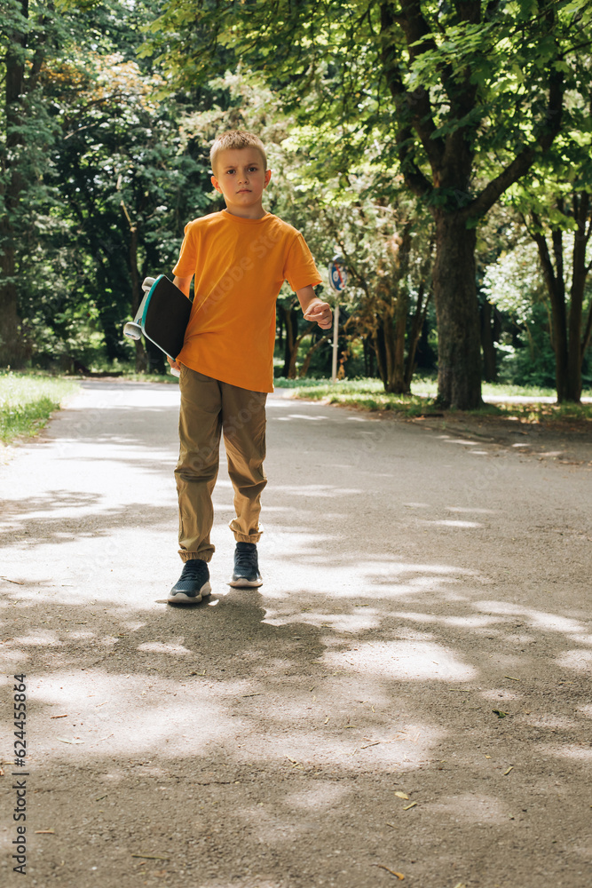 Outdoor activities for children. An upset young boy in sunglasses stay with a skateboard in a public park. Caucasian schoolboy on a walk. Front view Outdoor activities for children. An upset young boy in sunglasses stay with a skateboard in a public park. Caucasian schoolboy on a walk. Front view