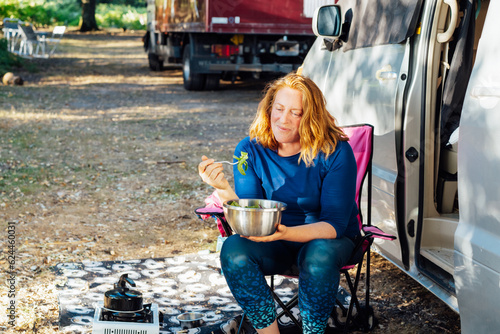 Relaxed Middle aged woman eating veggie salad outside camper van in the forest camp during road trip lunch. Enjoying nomad lifestyle, active weekend, vacation.