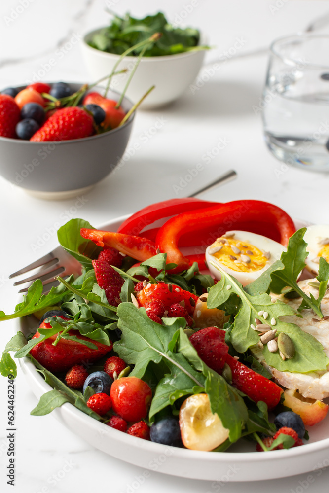 Healthy tasty breakfasts, a plate with boiled eggs and rice bread, salad of arugula and strawberries with cherries and blueberries, fresh peppers and seeds