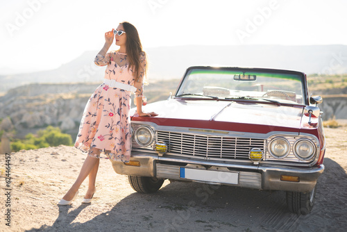 beautiful girl in a retro dress and sunglasses at sunrise happy posing on the background of the mountain landscape in Cappadocia near a red retro car