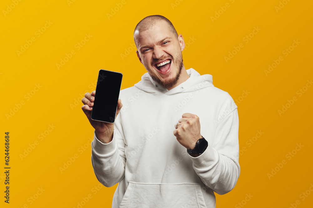 Excited young man screaming and showing empty screen smartphone while celebrating victory with raised fist, isolated over yellow background Excited young man screaming and showing empty screen smartphone while celebrating victory with raised fist, isolated over yellow background