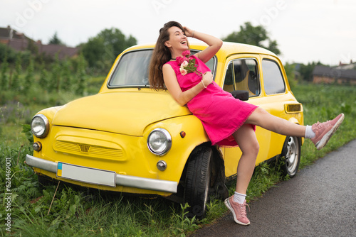 beautiful girl in a pink dress with a bouquet of red roses near a yellow retro car © Tsyb Oleh