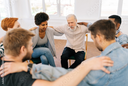 Top view of mature male therapist and group of multi-age patients putting hands on each others shoulders sitting in circle during group therapy session as symbol of support. Concept of mental health. © dikushin