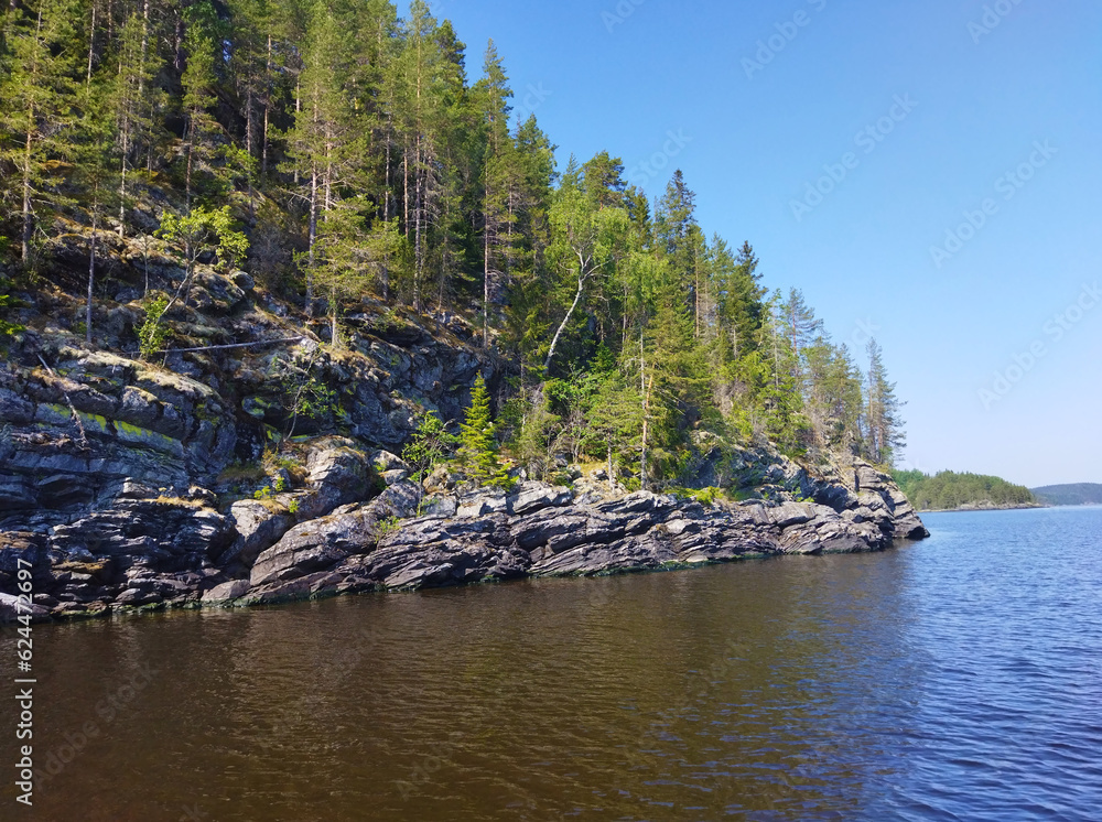 Rocky shore with large stones and trees reflected in calm water of northern lake. Beautiful summer nature of Ladoga lake. Karelia. Russia.
