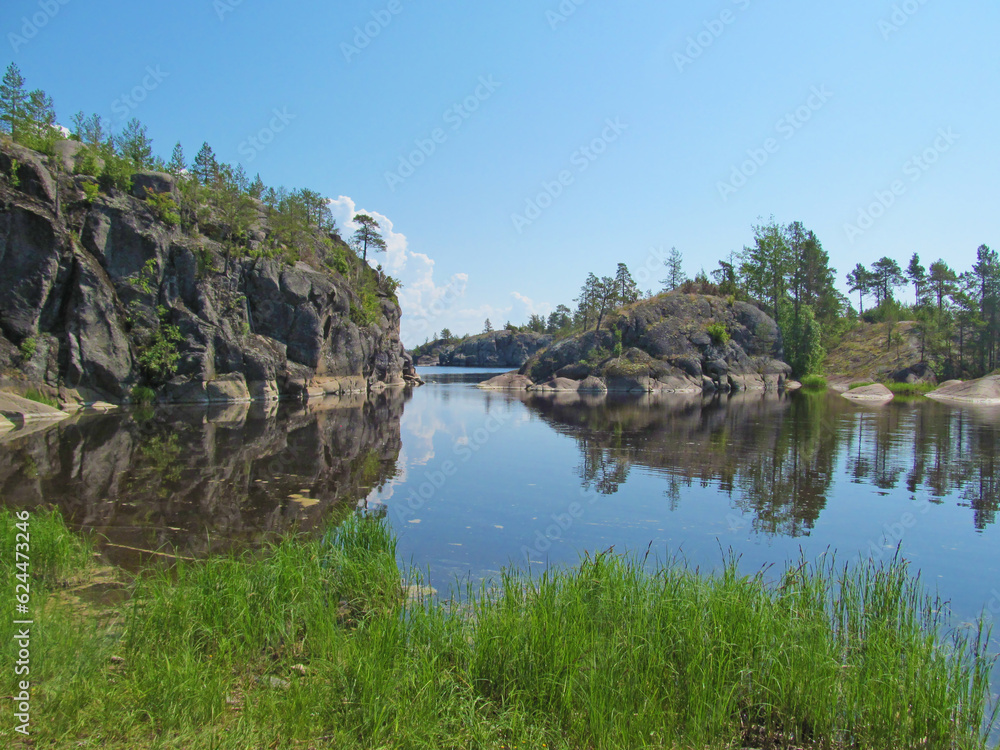 Rocky coast with large stones, trees and reflection in quiet water surface of northern lake. Northern nature and blue sky in summer. Ladoga lake, Karelia, Russia.