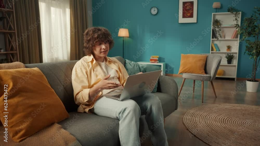 Portrait of a young curly guy in the living room on the sofa with a laptop on his lap. The guy communicates via video call, waves his hand in greeting, and nods his head. Portrait of a young curly guy in the living room on the sofa with a laptop on his lap. The guy communicates via video call, waves his hand in greeting, and nods his head.