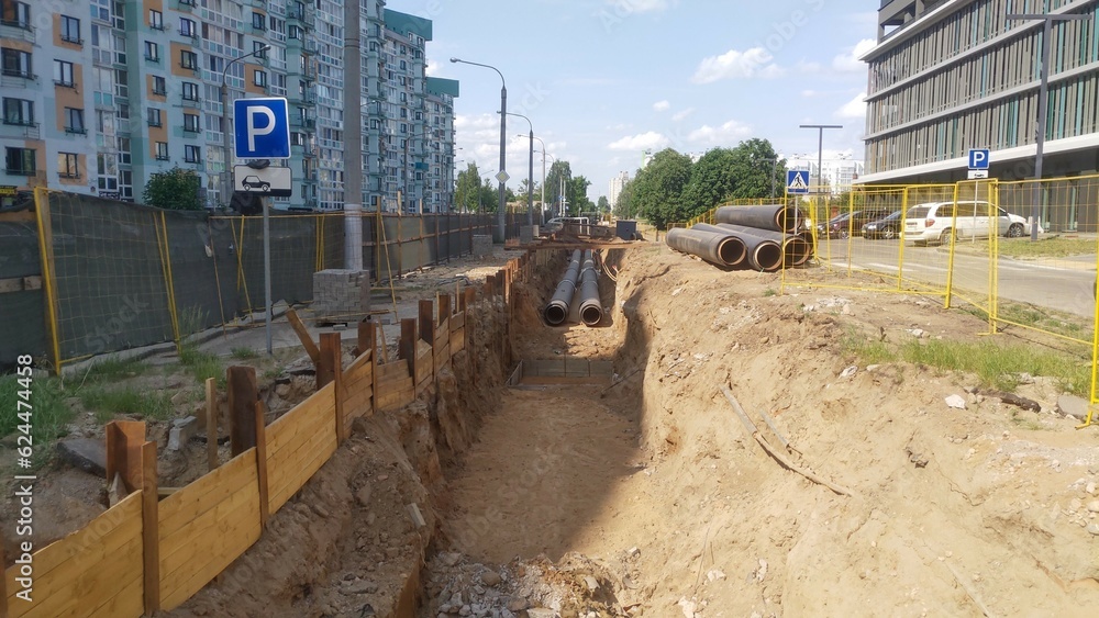 A trench was dug in a city street along the houses and asphalted roadway to replace cables, water and sewer pipes. For safety, the work site is enclosed with a metal fence. Sunny weather