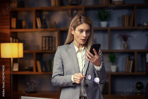 Surprised young woman makes big eyes opens, shocked with unprecedentedly notification or message, looking at mobile phone, working at home office. photo