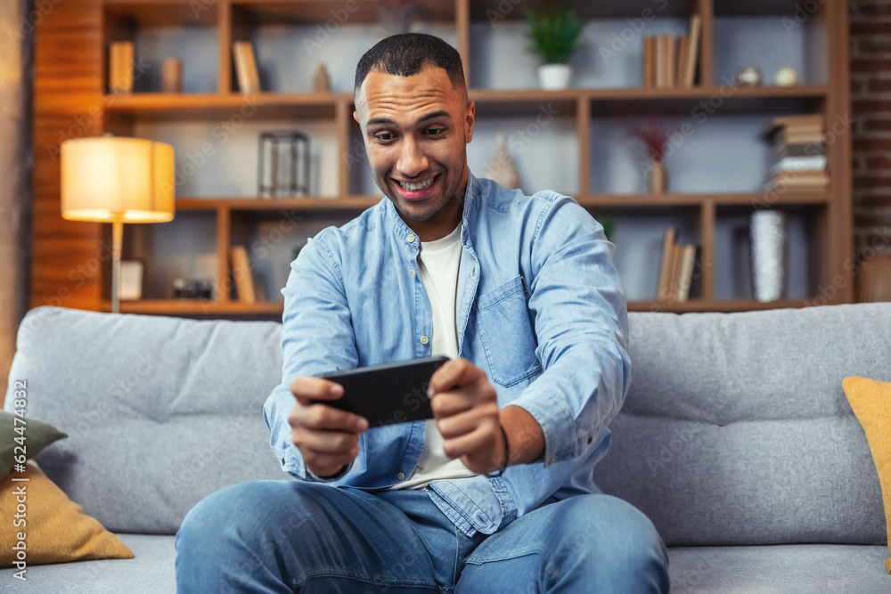 Excited happy young african american man gamer player using smartphone playing game in mobile videogame app or watching digital video stream holding cell phone Excited happy young african american man gamer player using smartphone playing game in mobile videogame app or watching digital video stream holding cell phone