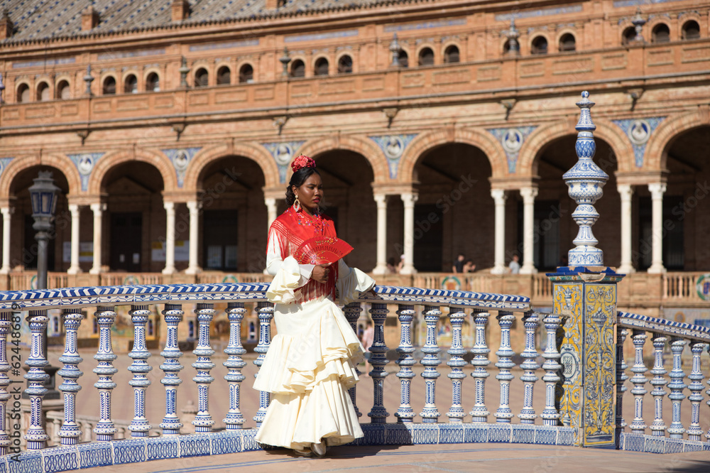 Young black woman dressed as a flamenco gypsy in a famous square in Seville, Spain. She wears a beige dress with ruffles and red shawl and red fan. Flamenco cultural heritage of humanity.