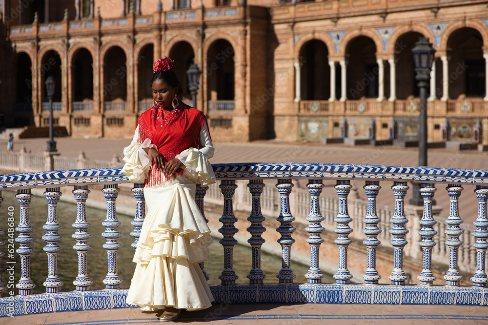 Young black woman dressed as a flamenco gypsy in a square in Seville, Spain. She wears a beige dress with ruffles and a red shawl with flowers. Flamenco cultural heritage of humanity. Dance concept.