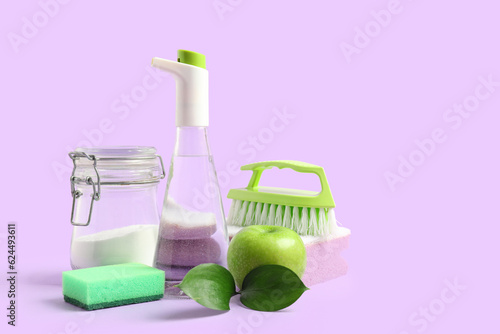 Bottle of vinegar, baking soda, sponges, brush and apple on lilac background