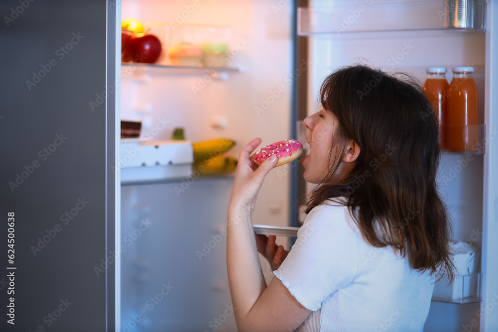 Young woman eating tasty doughnut near open fridge in kitchen at night Young woman eating tasty doughnut near open fridge in kitchen at night
