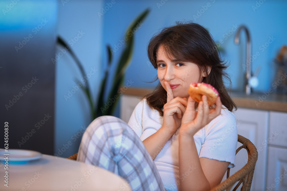 Young woman eating tasty doughnut in kitchen at night Young woman eating tasty doughnut in kitchen at night