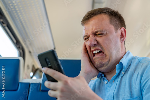 Disgusted and overwhelmed man stares at screen of smartphone with confuse, bad joke or inappropriate content. Sad millennial guy looks at phone screen in surprise, sits in commuter train.